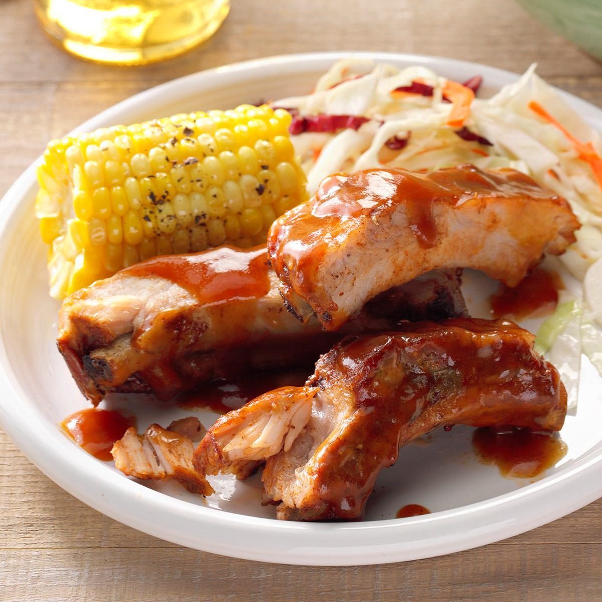 Barbecue ribs are coated in sauce, served with corn on the cob and coleslaw on a white plate, placed on a wooden table.