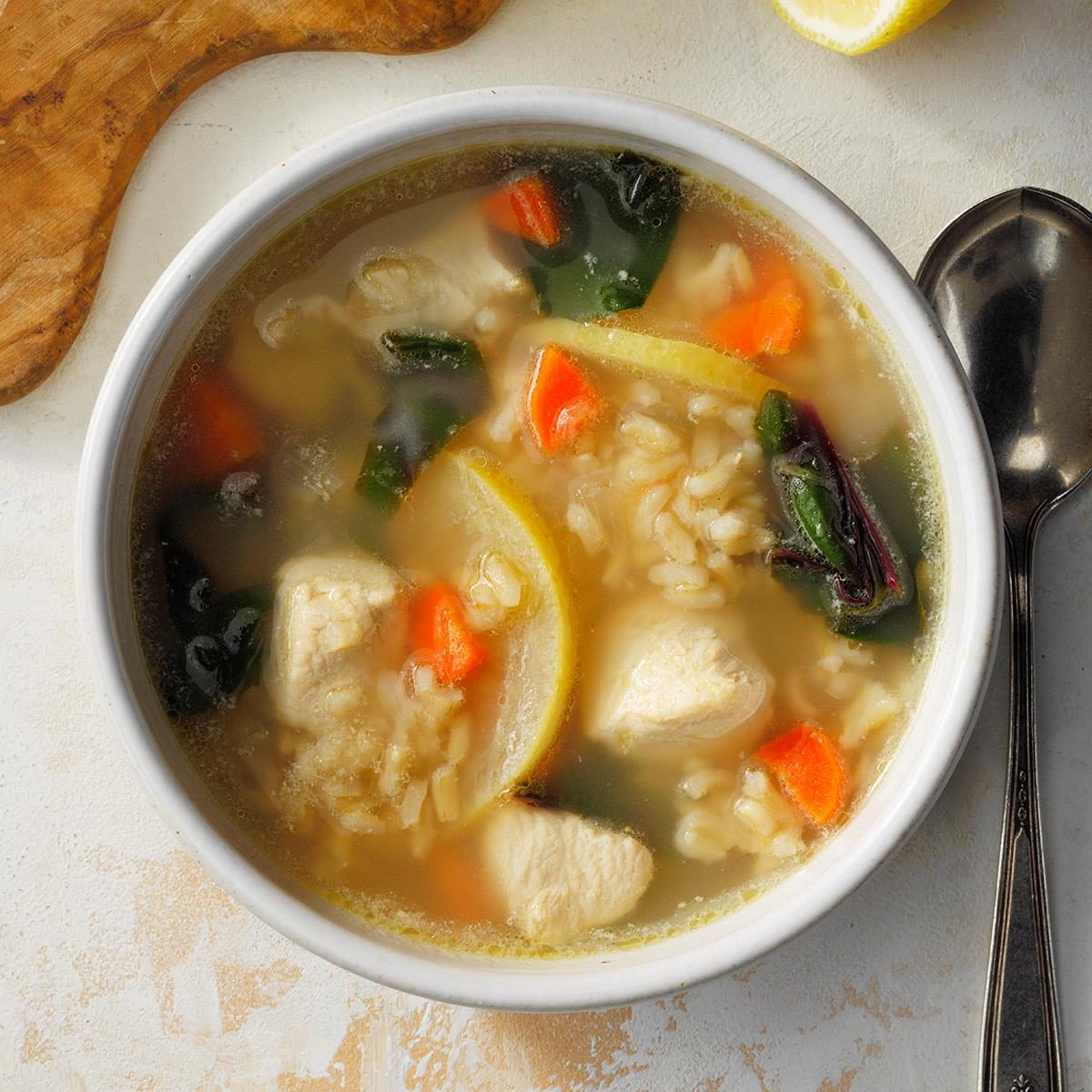 Bowl contains soup with chicken, rice, carrots, lemon slices, and greens; spoon beside on a light surface.