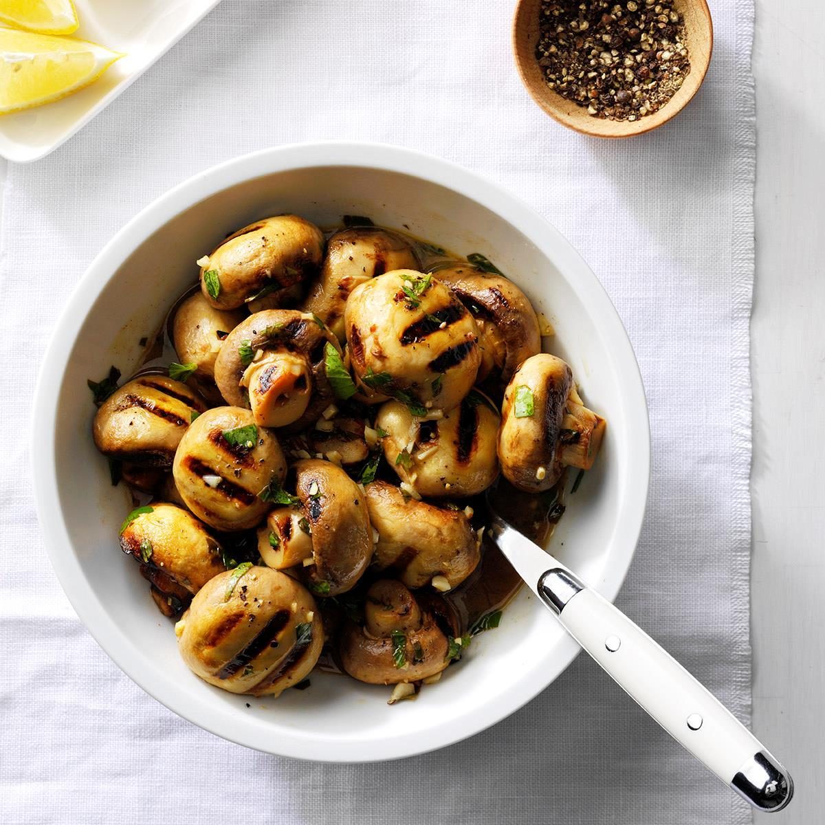Grilled mushrooms sit in a white bowl on a tablecloth, accompanied by sliced lemons and a bowl of pepper.