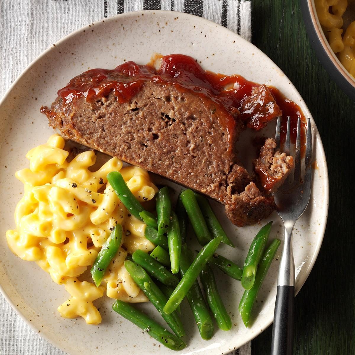 Plate holds meatloaf with tomato sauce, macaroni and cheese, and green beans; a fork rests beside the meatloaf, setting on a dark green table.