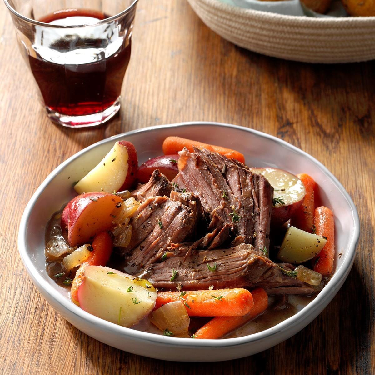 Plate of pot roast with carrots and potatoes on a wooden table beside a glass of red beverage.