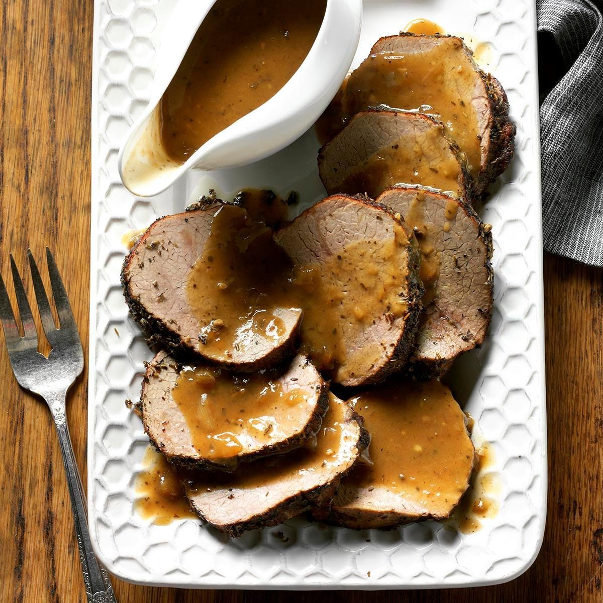 Slices of roast beef are drizzled with gravy on a white platter, accompanied by a gravy boat, on a wooden table.