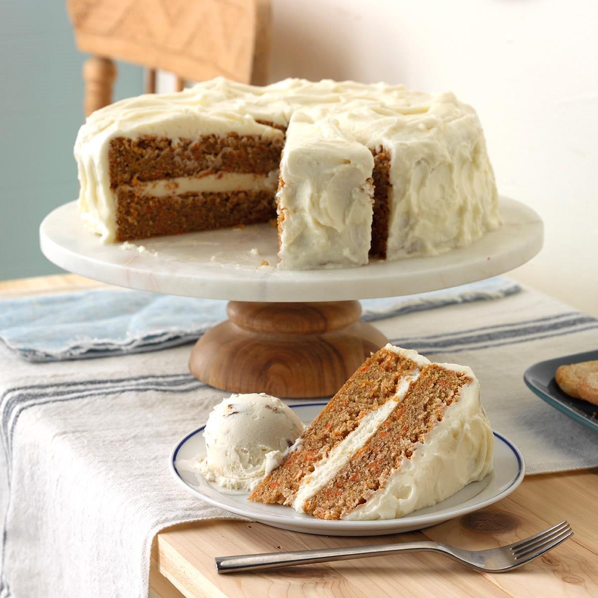 Carrot cake with white frosting sits on a wooden stand; a slice with ice cream is placed on a nearby plate on a table.