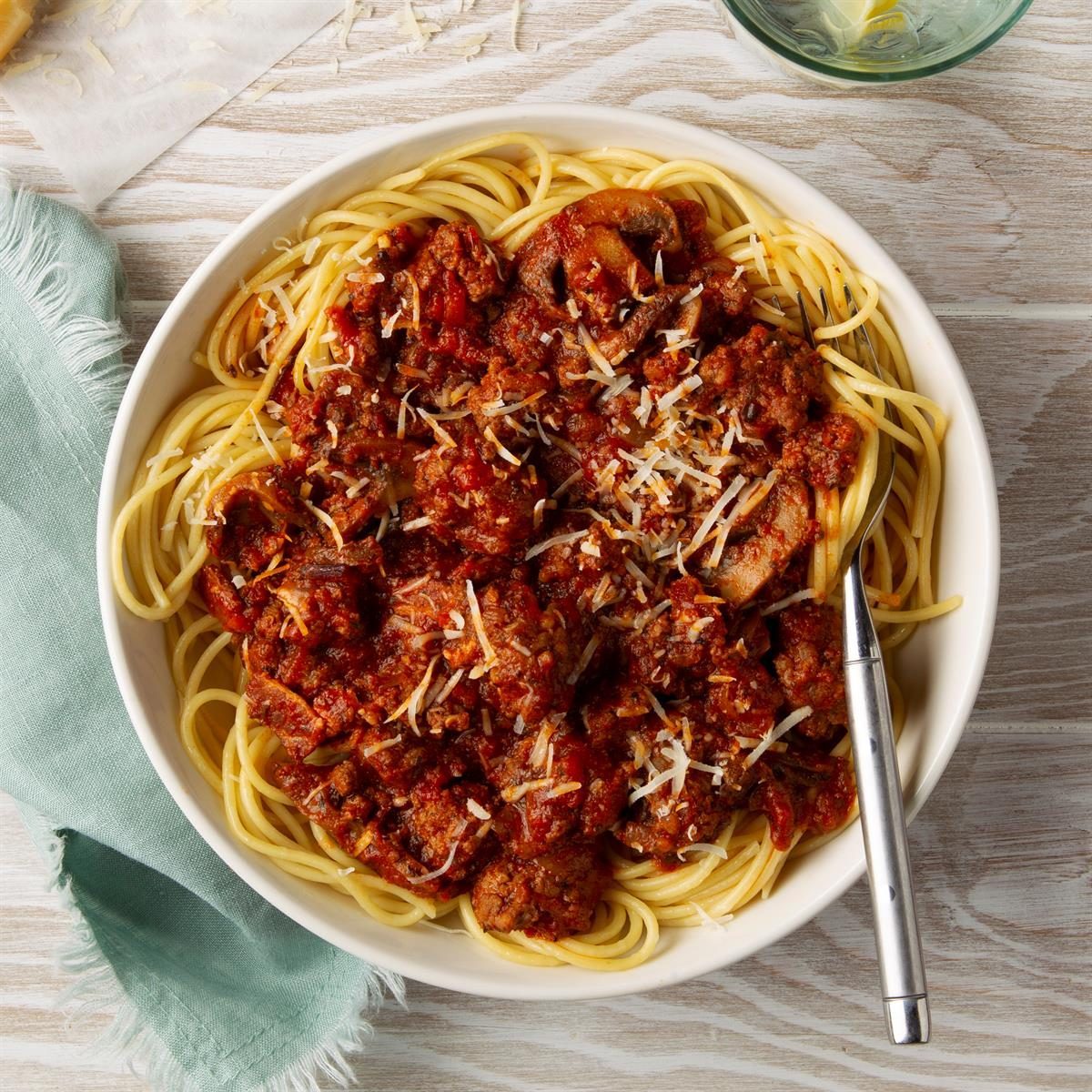 Spaghetti topped with meat sauce and cheese rests in a white bowl, set on a wooden table with a green fabric napkin and a fork.