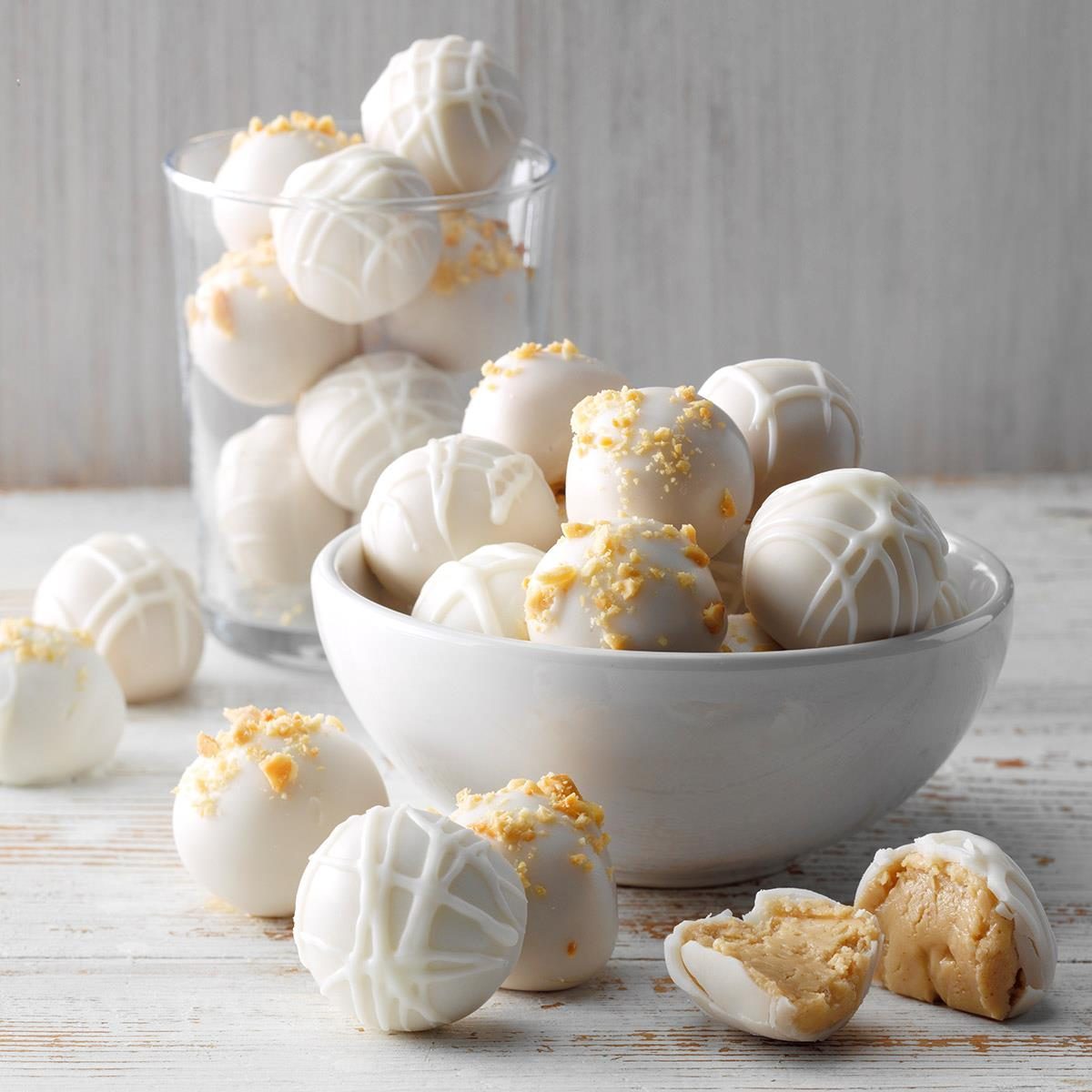 White chocolate truffles resting in and around a bowl; some decorated with drizzle, others with crumbs, set on a rustic wooden table.