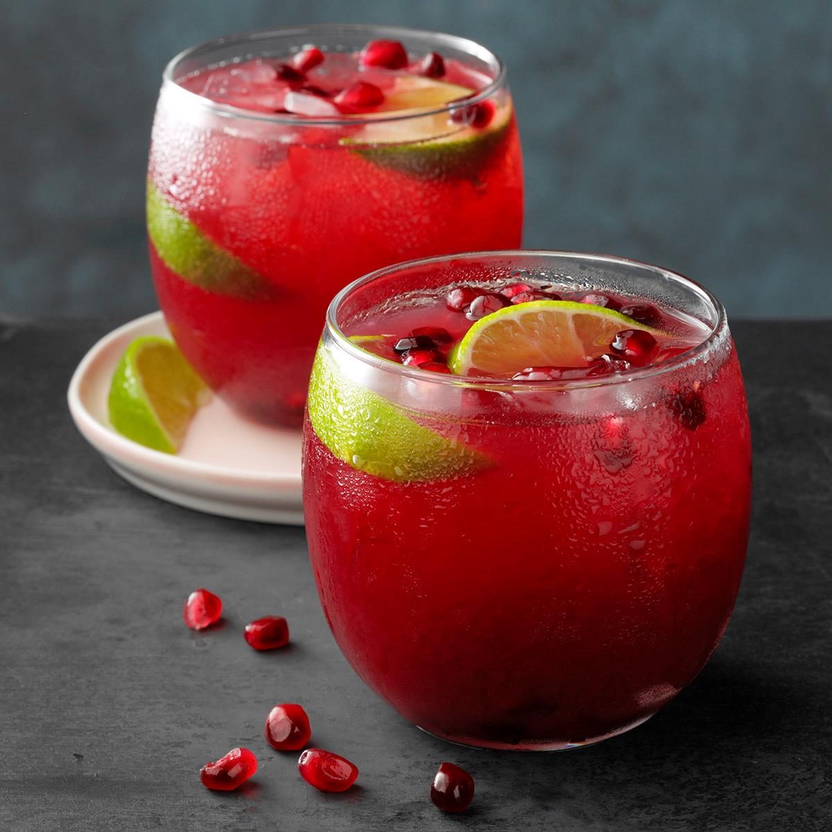 Red beverage sits in rounded glasses, accented with lime wedges and pomegranate seeds, on a dark surface with a blurred background.
