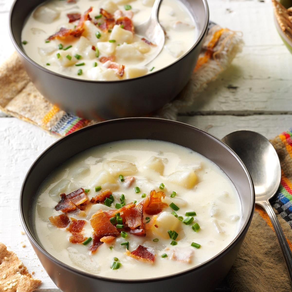 Bowls of creamy potato soup, garnished with bacon and chives, sit on a table with a colorful fabric napkin and spoon nearby.