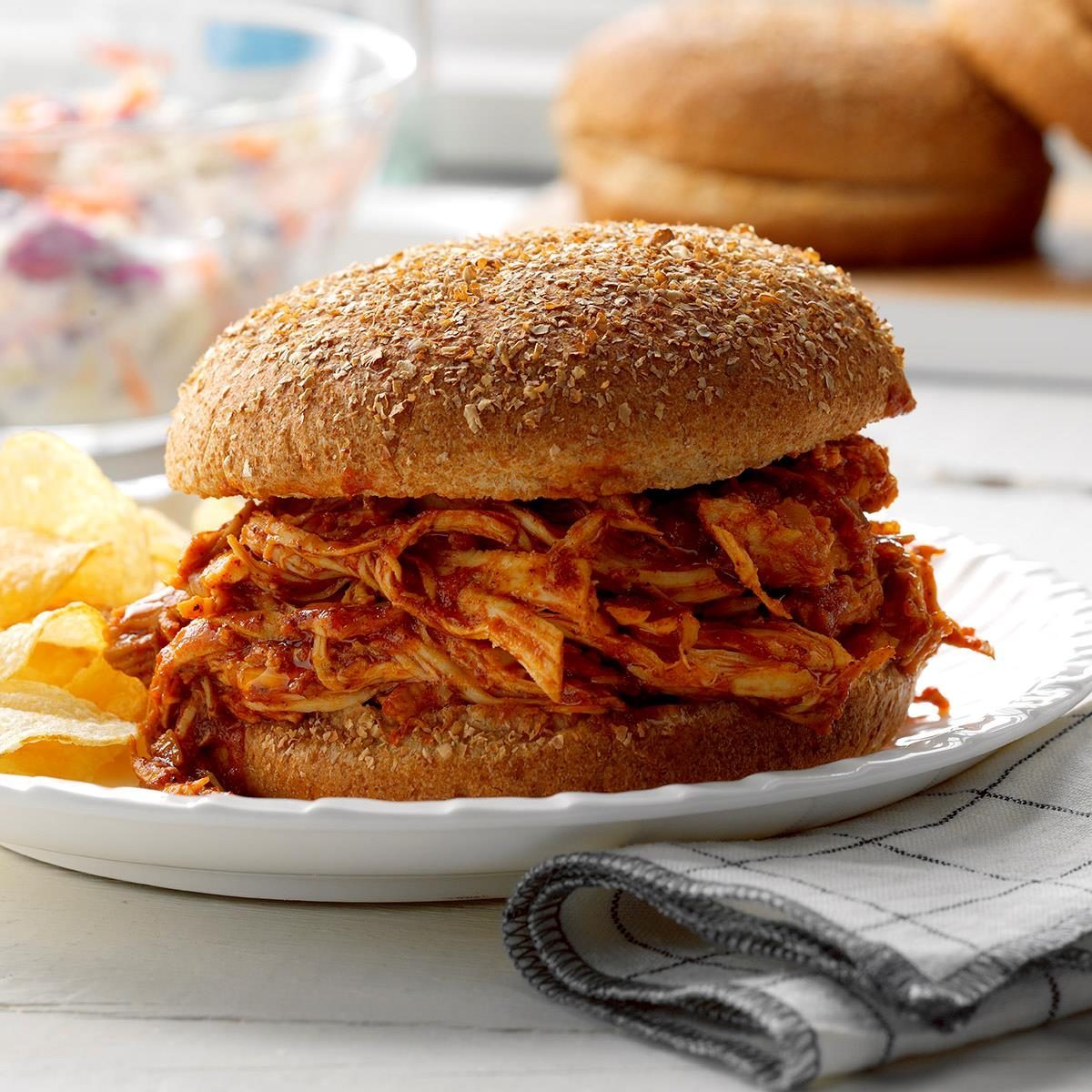 A sandwich overflowing with saucy shredded chicken rests on a white plate, accompanied by potato chips, with a blurred coleslaw background.