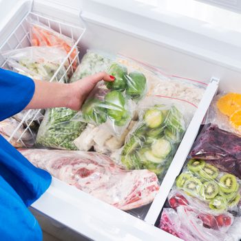 Hand holds a bag of green peppers in an open freezer filled with various frozen vegetables, meat, and fruits in plastic bags.