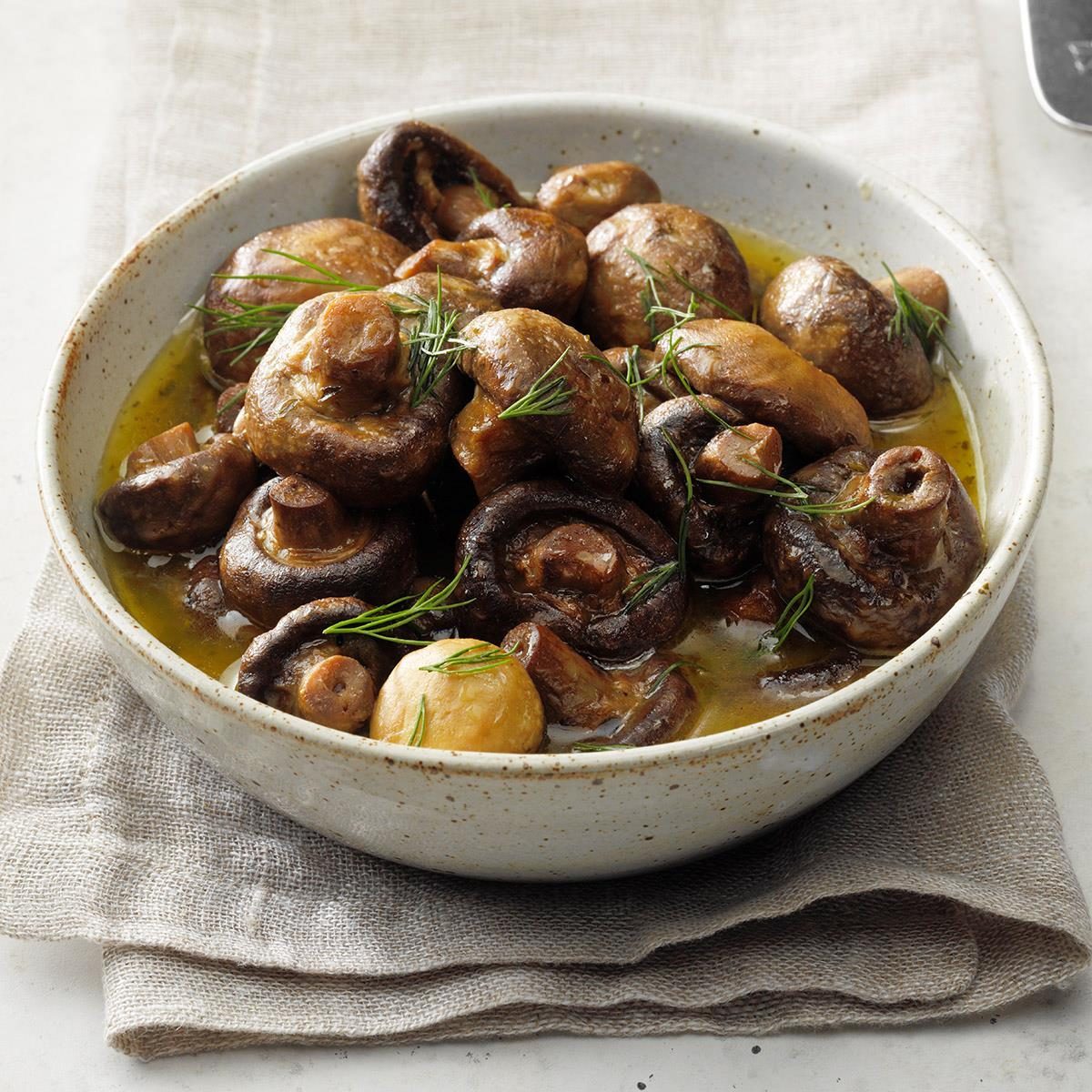 Mushrooms rest in a bowl, garnished with herbs, and surrounded by a light sauce, set on a textured cloth.