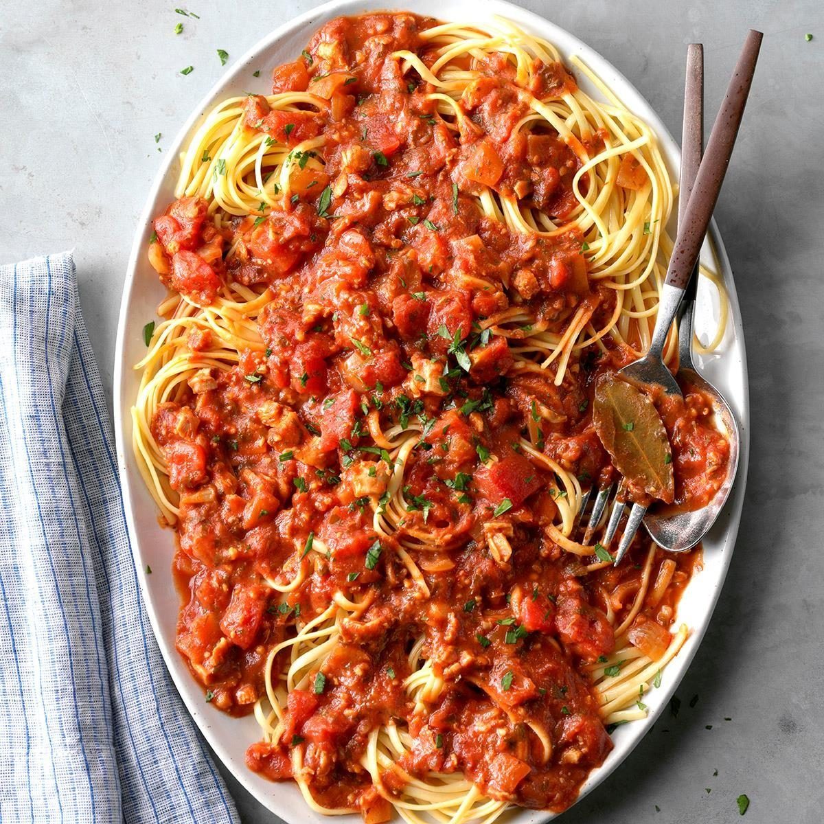Spaghetti sits covered in rich tomato sauce on a white plate, garnished with herbs and accompanied by a fork and spoon.