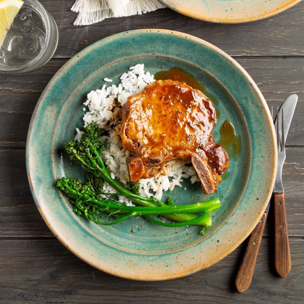 A glazed pork chop sits on rice with broccolini, served on a turquoise plate, accompanied by utensils on a wooden table.