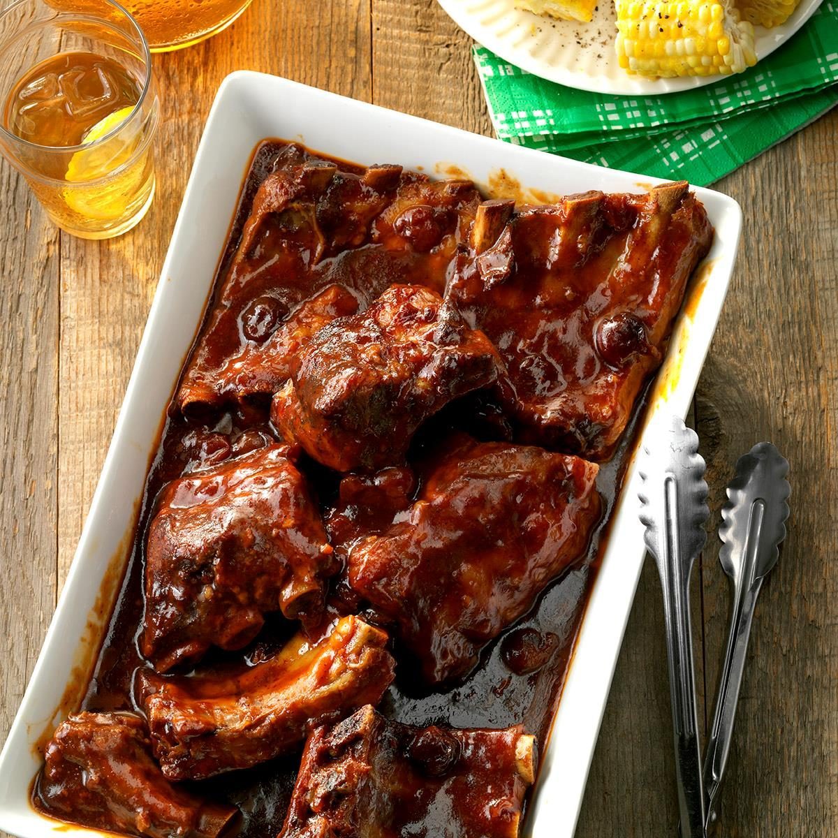 Barbecue ribs sit in a rectangular dish, covered in sauce, on a wooden table with a drink and corn-on-the-cob on a plate nearby.