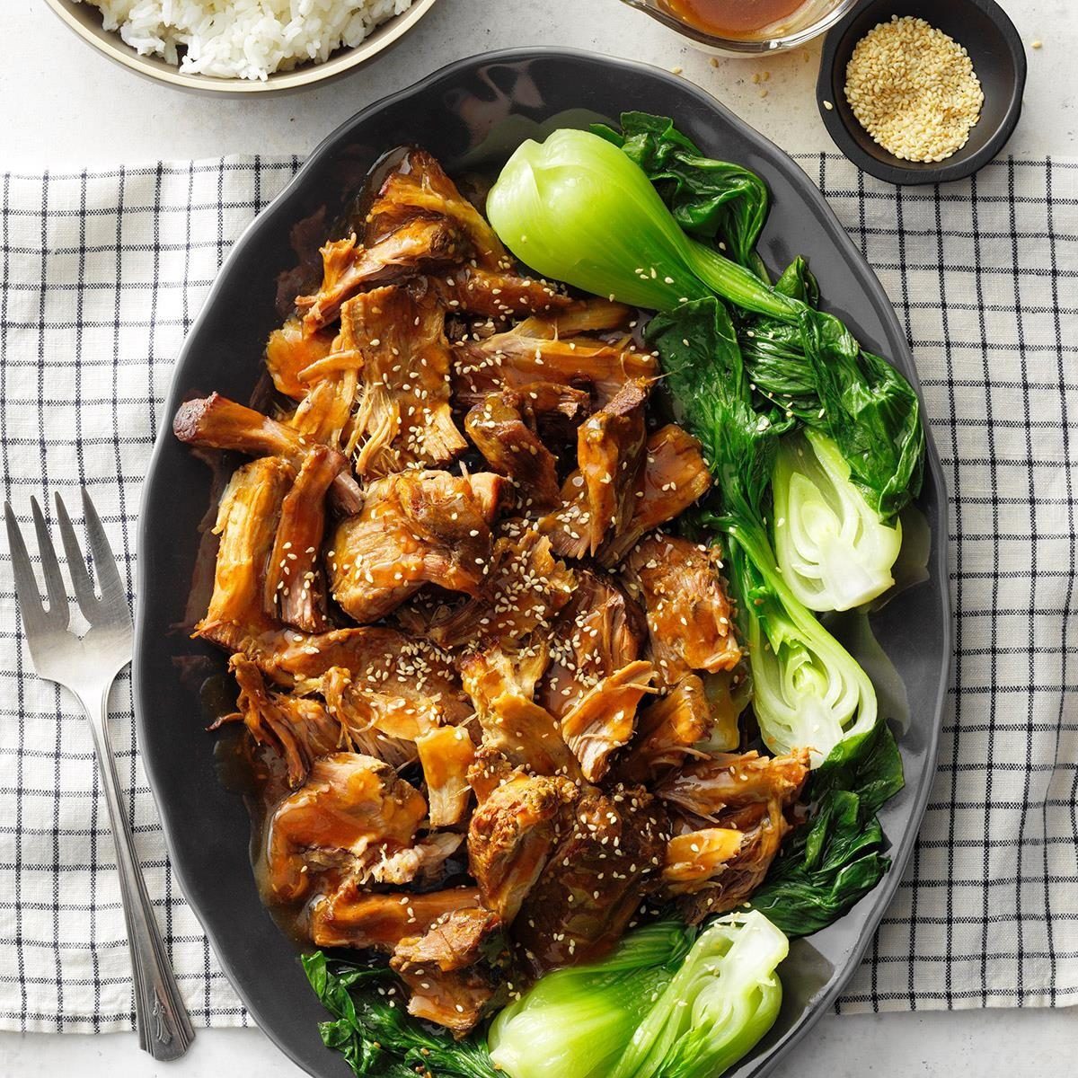 Shredded meat topped with sesame seeds, surrounded by bok choy on a black plate, placed on a checkered cloth with rice and sauce nearby.