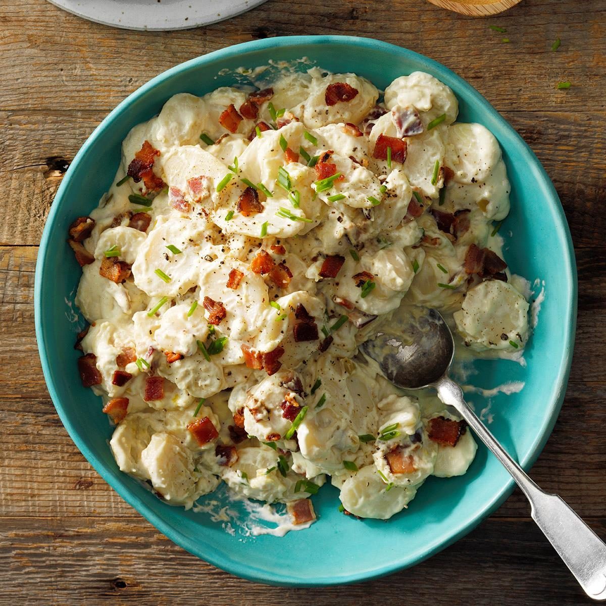 Potato salad topped with bacon and chives rests in a turquoise bowl, surrounded by a rustic wooden table.