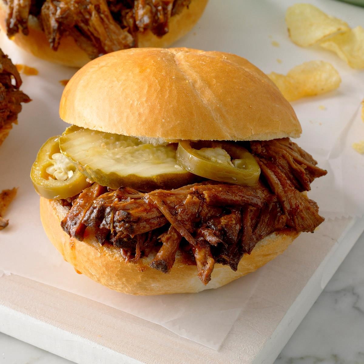 Sandwich filled with shredded meat, pickles, and jalapeÃ±os resting on a white surface, accompanied by potato chips in the background.