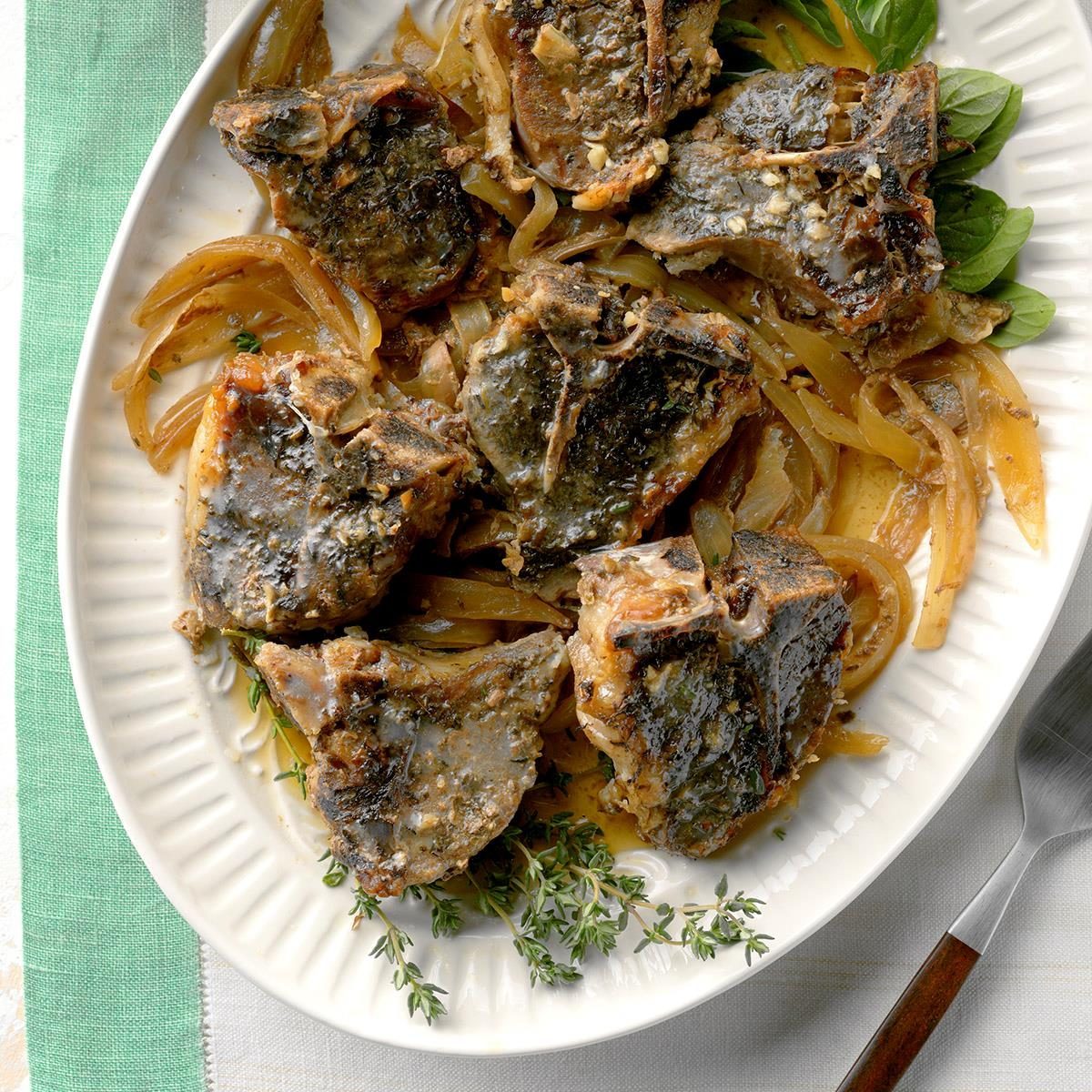 Lamb chops rest on sautÃ©ed onions, garnished with fresh thyme and basil, served on a white oval plate over a green and white tablecloth.