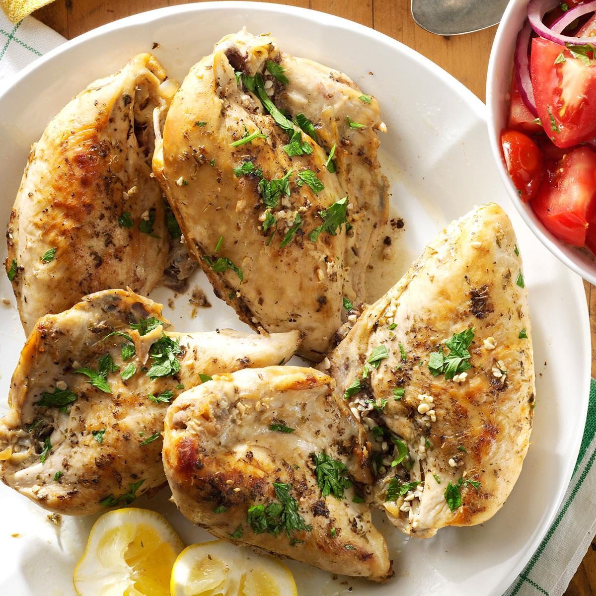 Chicken breasts garnished with herbs are arranged on a plate, accompanied by lemon slices and a bowl of tomato and onion salad nearby.