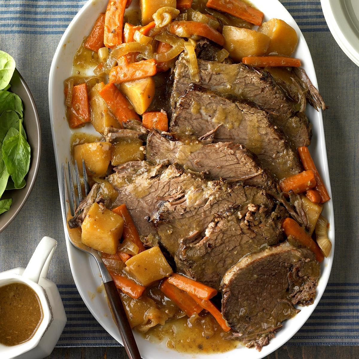 Slices of roast beef sit with carrots and potatoes on a white platter, surrounded by gravy, atop a striped cloth next to a dish with spinach.