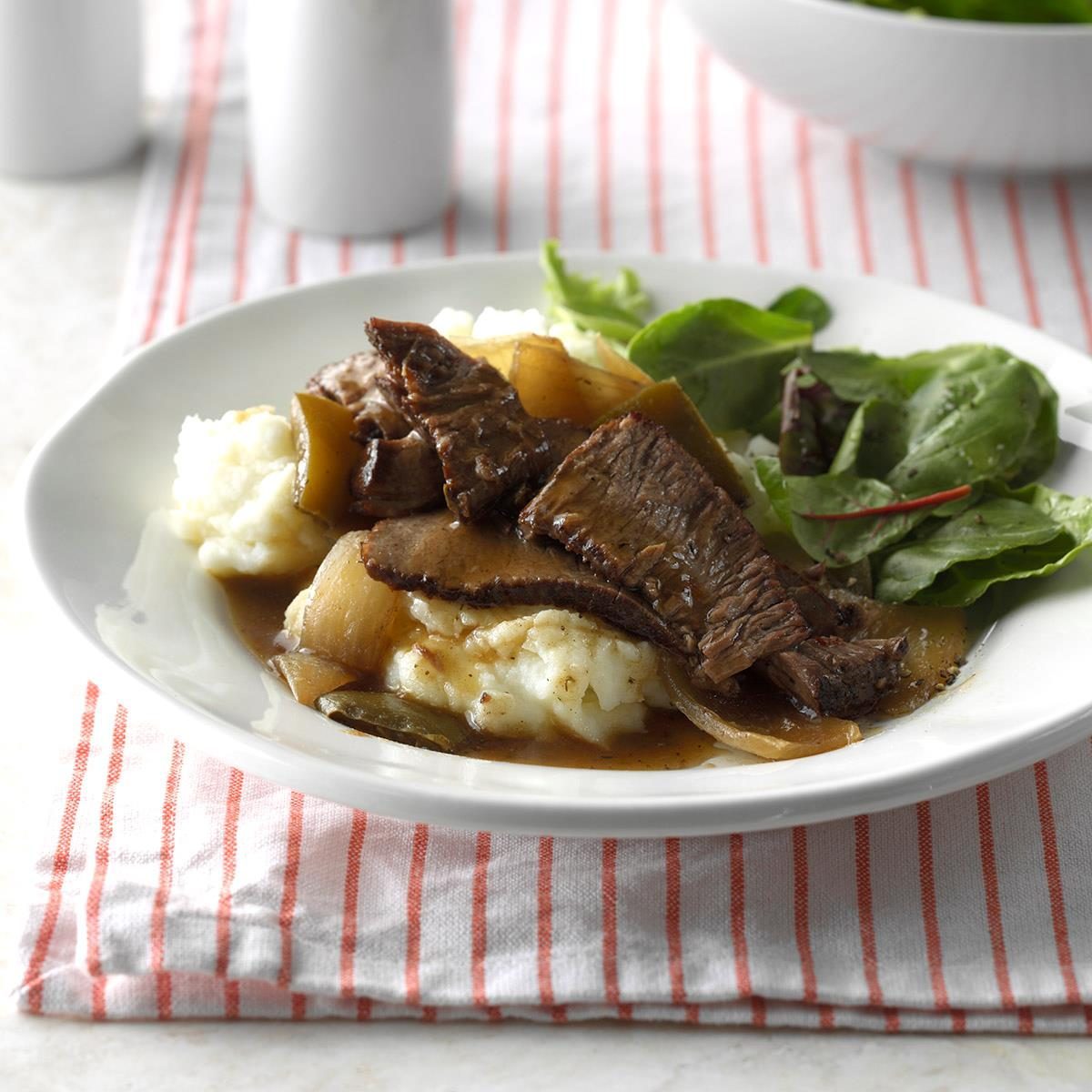 Beef slices resting on mashed potatoes with gravy, accompanied by fresh greens on a white plate, placed on a red-striped cloth.