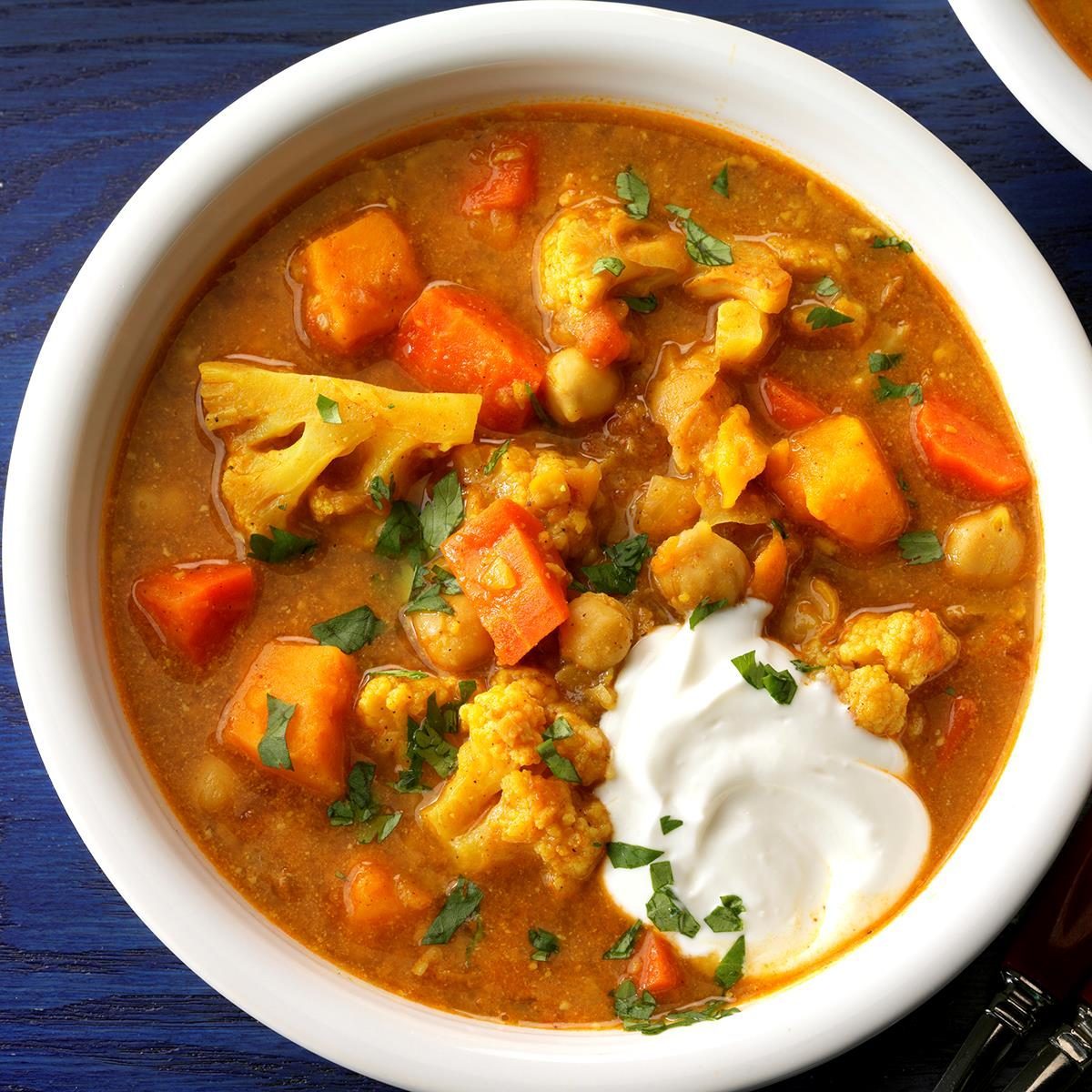 Bowl holds vegetable curry with chickpeas and cauliflower, garnished with cilantro and a dollop of yogurt, set on a dark blue surface.