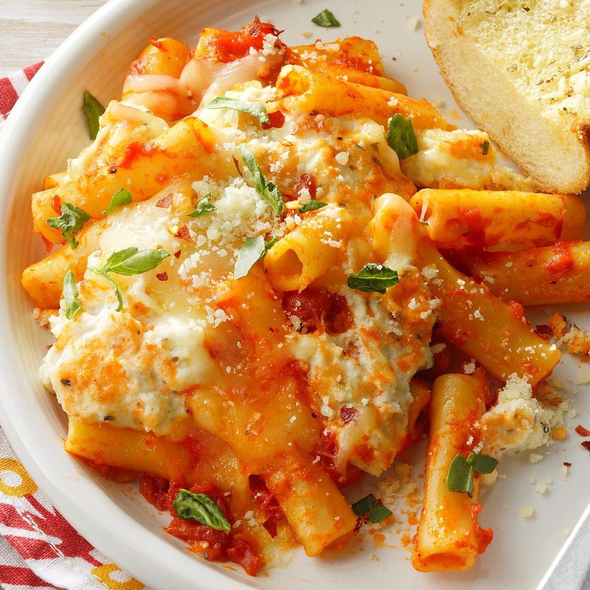 Baked ziti topped with melted cheese and basil, served with a slice of garlic bread on a white plate.