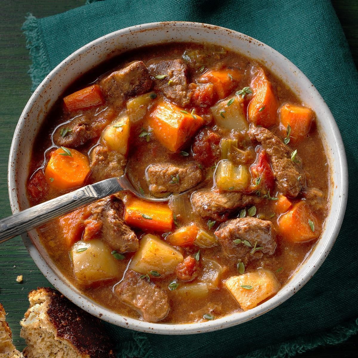 Hearty stew fills a bowl, showcasing chunks of beef and vegetables in savory broth, set on a dark table with a green cloth.