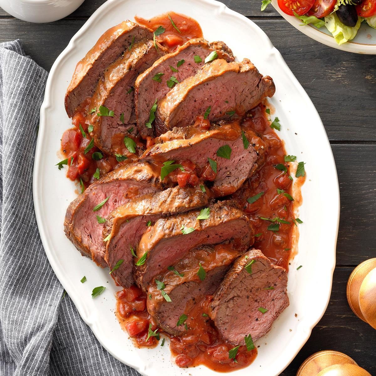 Sliced beef roast drizzled with tomato sauce, garnished with herbs on a white platter, placed on a dark wooden table alongside a striped cloth.