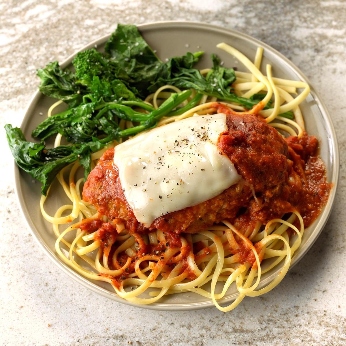 A plate holds chicken Parmesan with melted cheese, resting on spaghetti, accompanied by sautÃ©ed spinach, presented on a speckled countertop.
