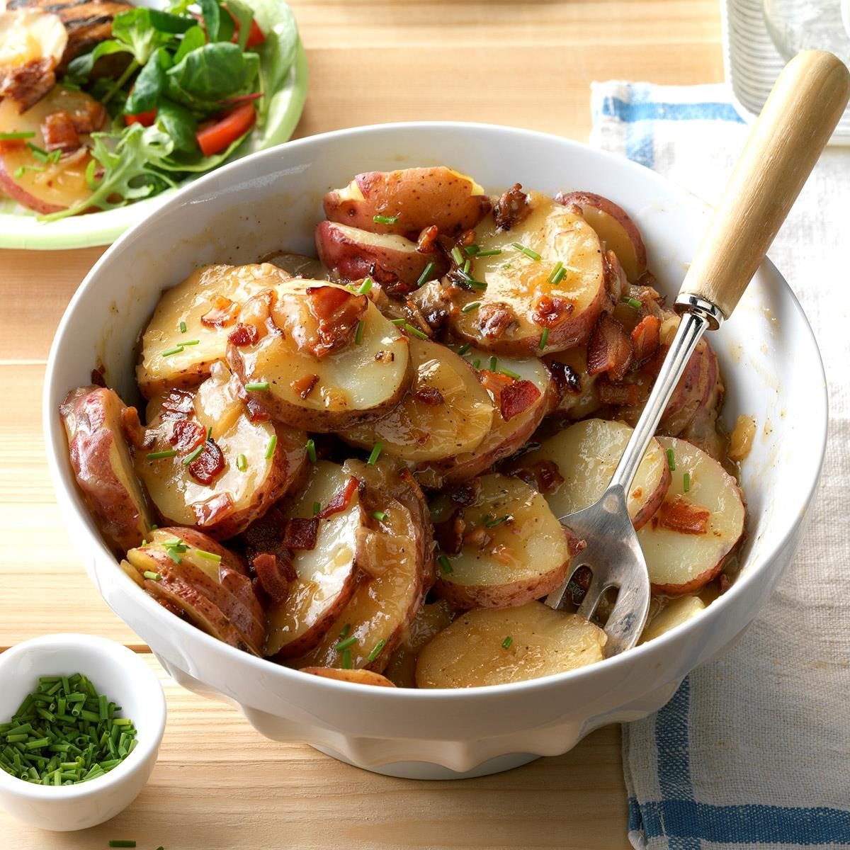 Bowl contains sliced potatoes with bacon and chives, accompanied by a fork; salad sits nearby on a wooden table.