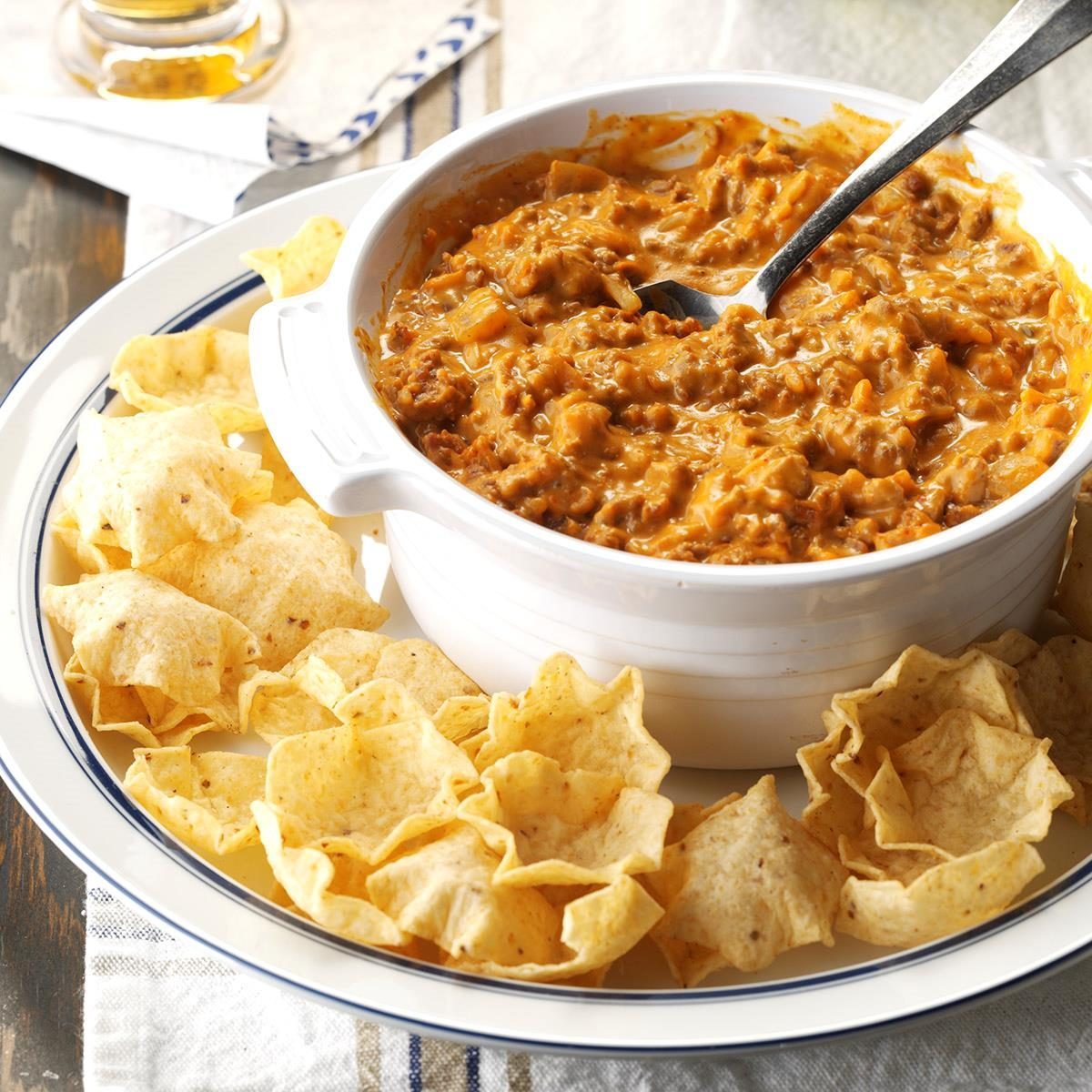 Cheesy dip sits in a white bowl with a spoon, surrounded by tortilla chips on a plate, atop a table with a striped napkin.