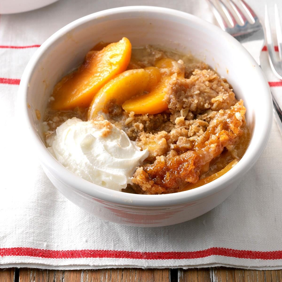 Bowl contains peach cobbler with whipped cream; placed on a red-striped white napkin beside a fork on a wooden table.