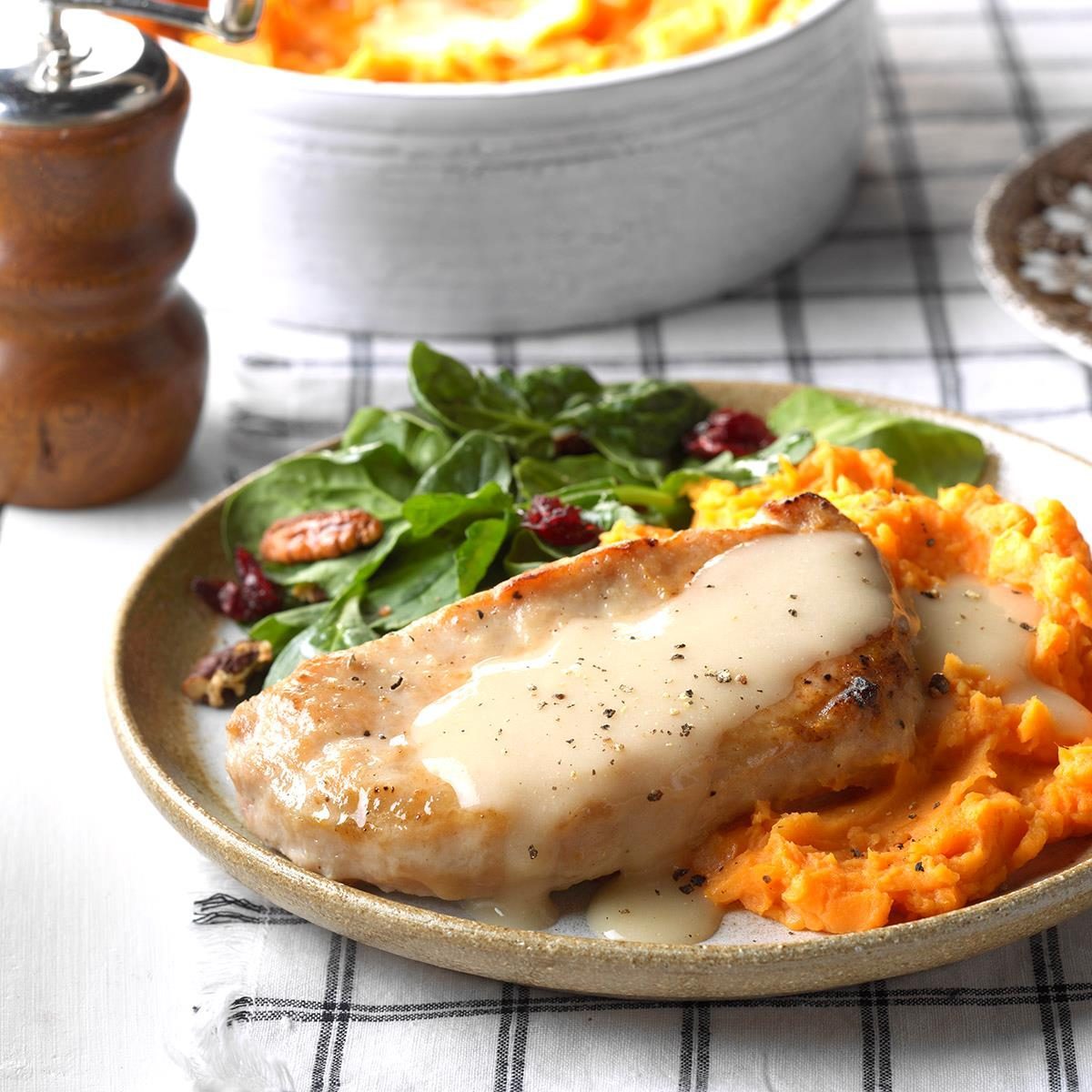 Pork chop sits on mashed sweet potatoes, drizzled with creamy sauce, accompanied by mixed greens with nuts and cranberries on a plaid-patterned tablecloth.