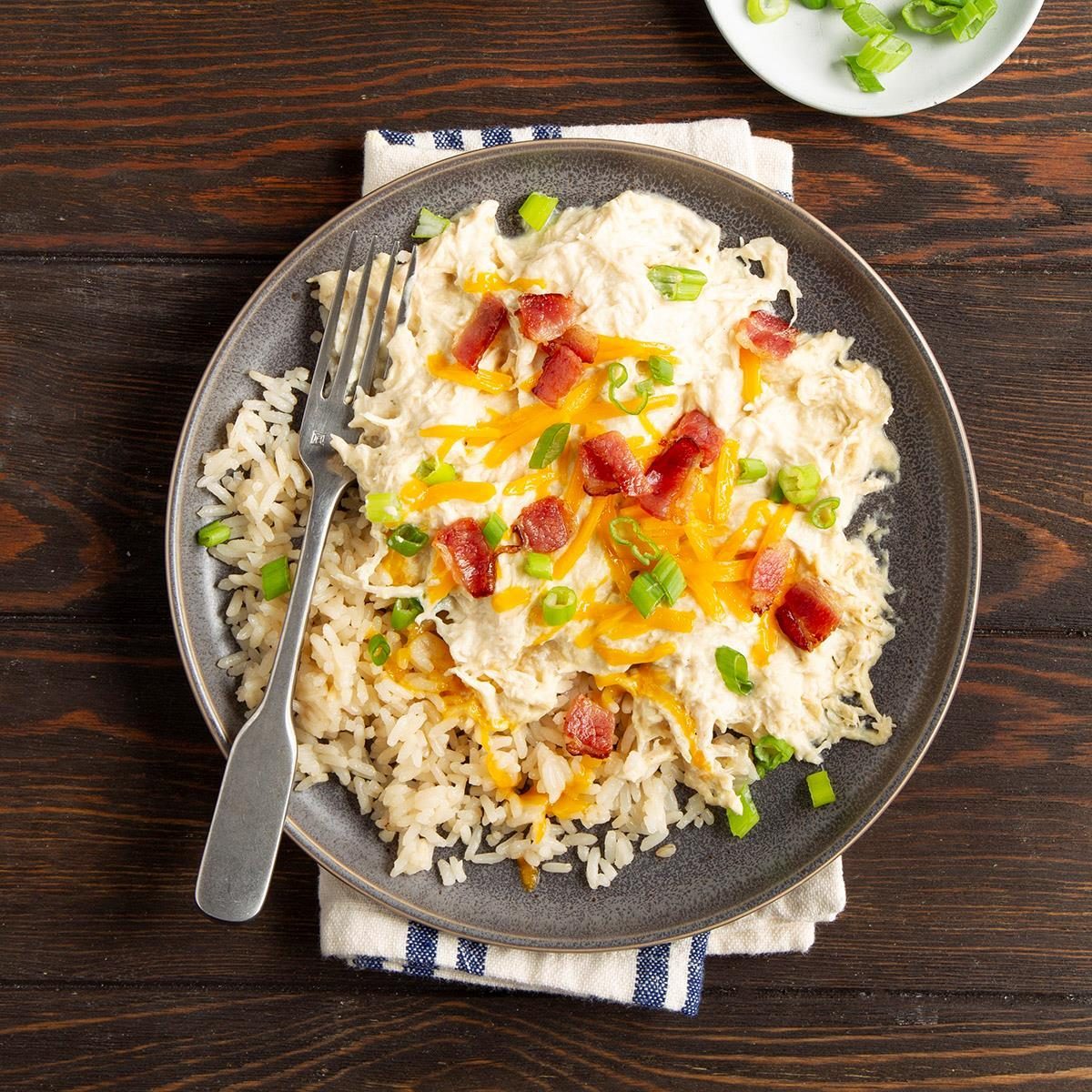 A plate holds chicken, rice, cheese, bacon, and green onions, with a fork positioned on a wooden table background.