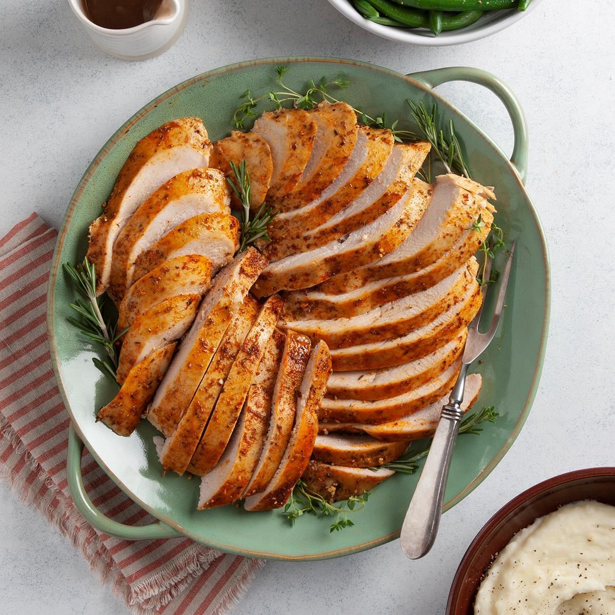 Sliced turkey breast arranged on a green platter with herbs, accompanied by mashed potatoes, green beans, and a small jug of gravy.