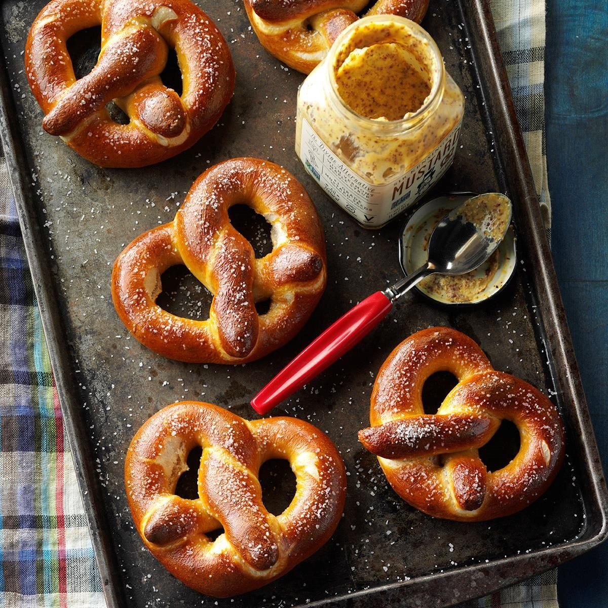 Fresh pretzels sit on a baking tray beside an open jar of mustard with a red-handled spoon, atop a checkered cloth.