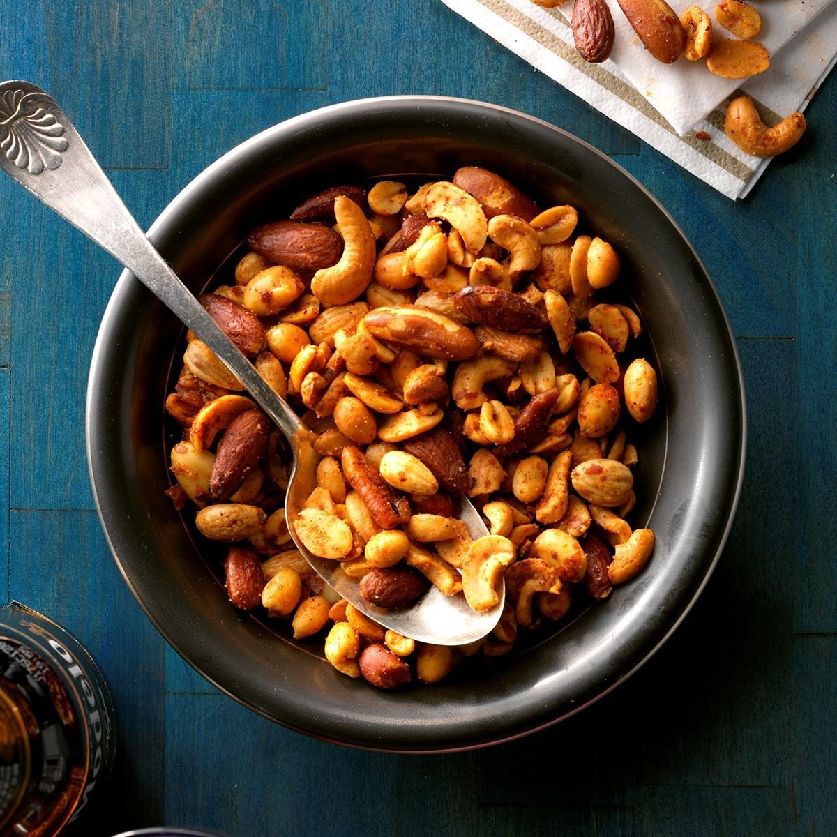 Bowl holds mixed nuts with spoon; scattered nuts on napkin nearby on a blue surface.