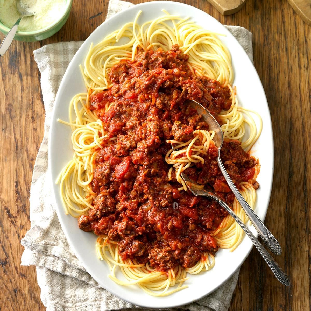 Spaghetti covered in meat sauce sits on a white oval plate with serving utensils, placed on a wooden table with a napkin.
