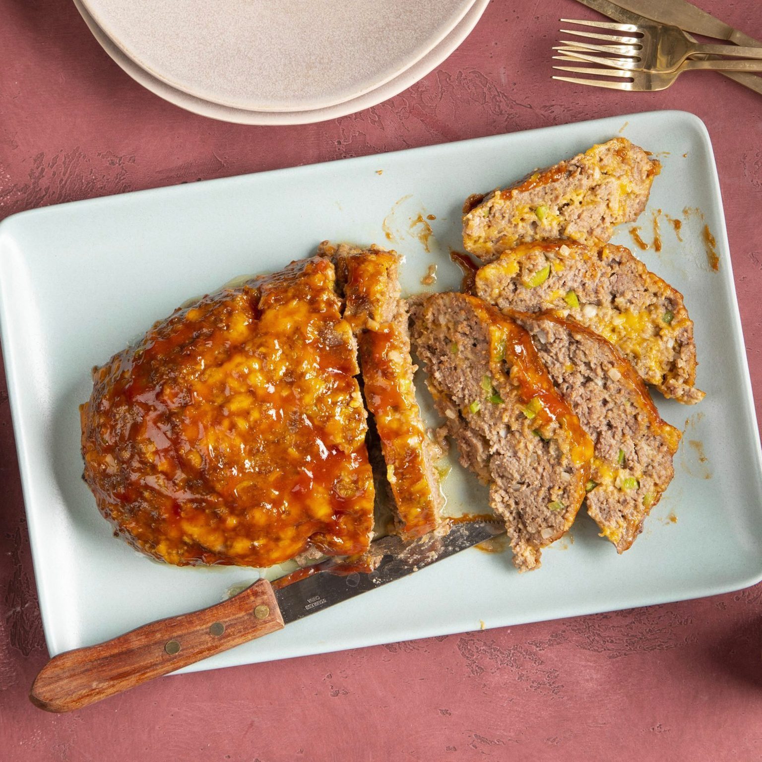 Meatloaf sliced on a pale blue platter with shiny glaze, accompanied by a wooden-handled knife; set on a textured pink surface.