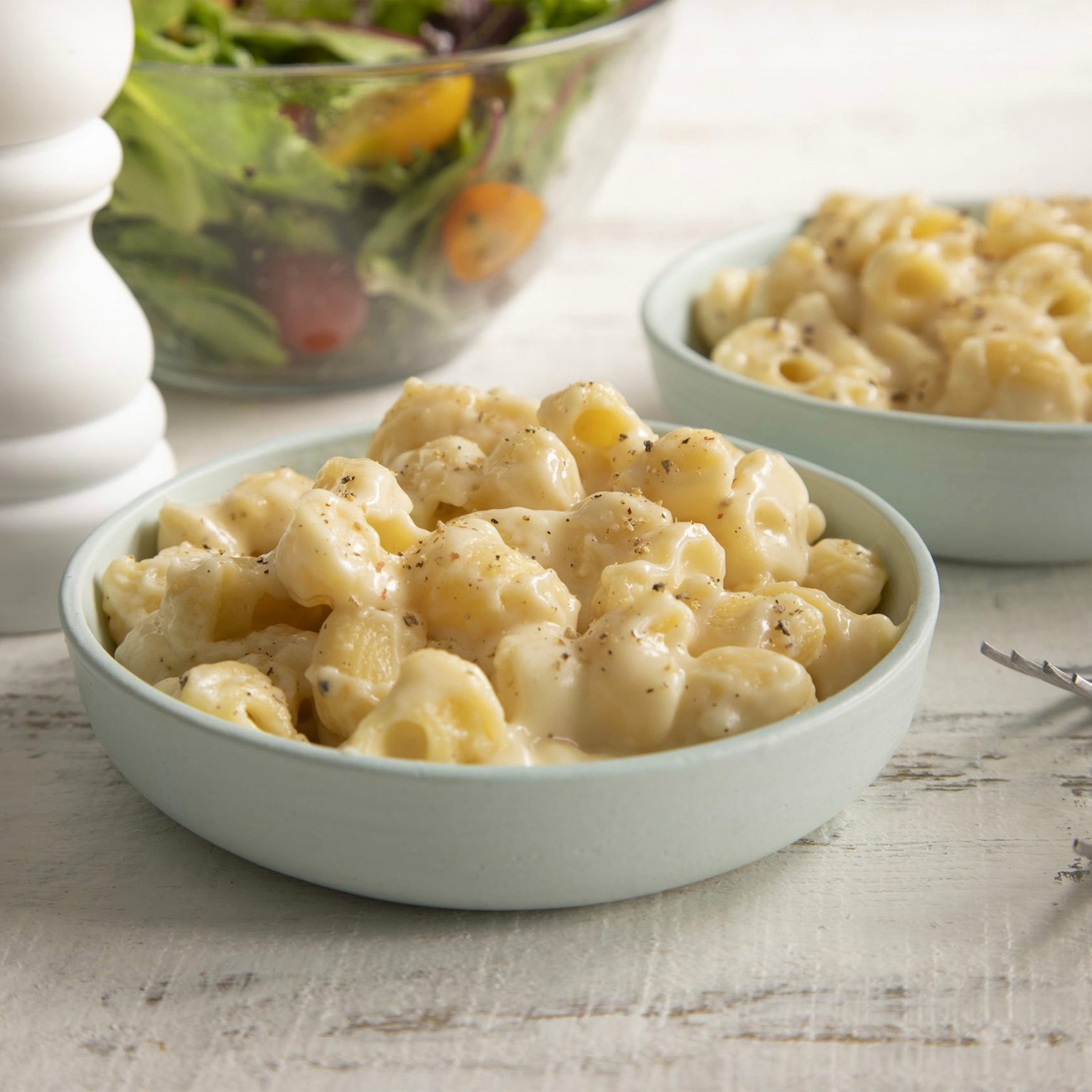 Bowls of creamy macaroni and cheese sit on a white table, with a fresh salad and a pepper grinder in the background.