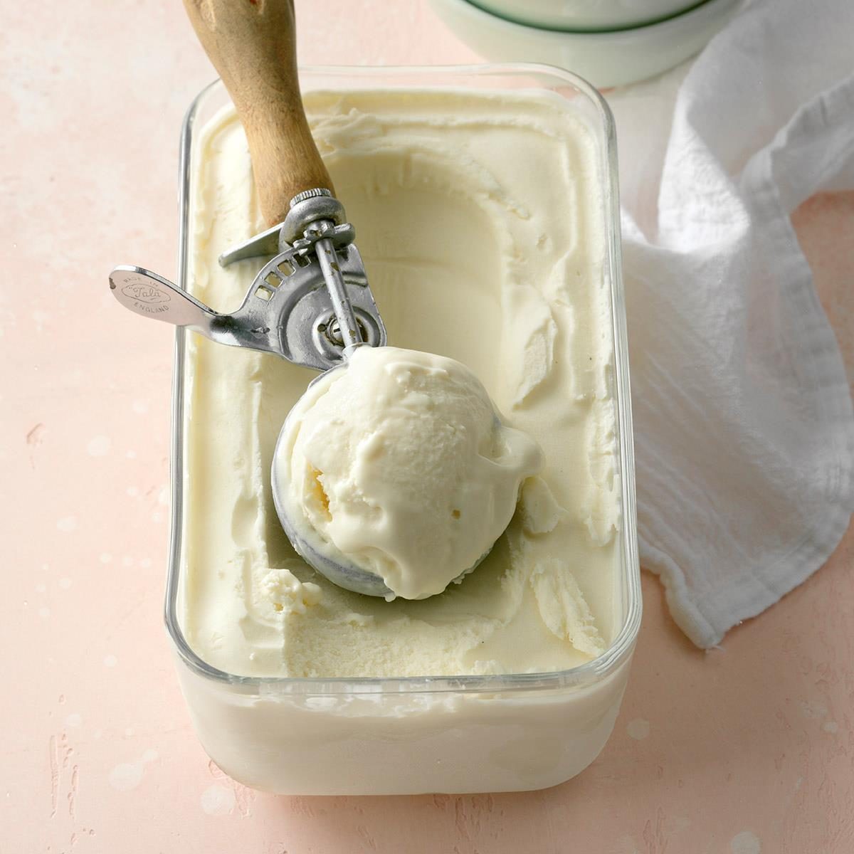 Ice cream scoop holding vanilla ice cream in a rectangular container, placed on a light pink surface with a white cloth nearby.