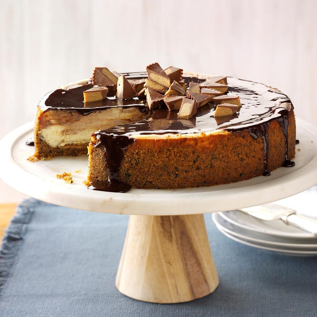 Cake topped with chocolate glaze and chopped peanut butter cups, placed on a wooden pedestal, against a simple background with no visible text.