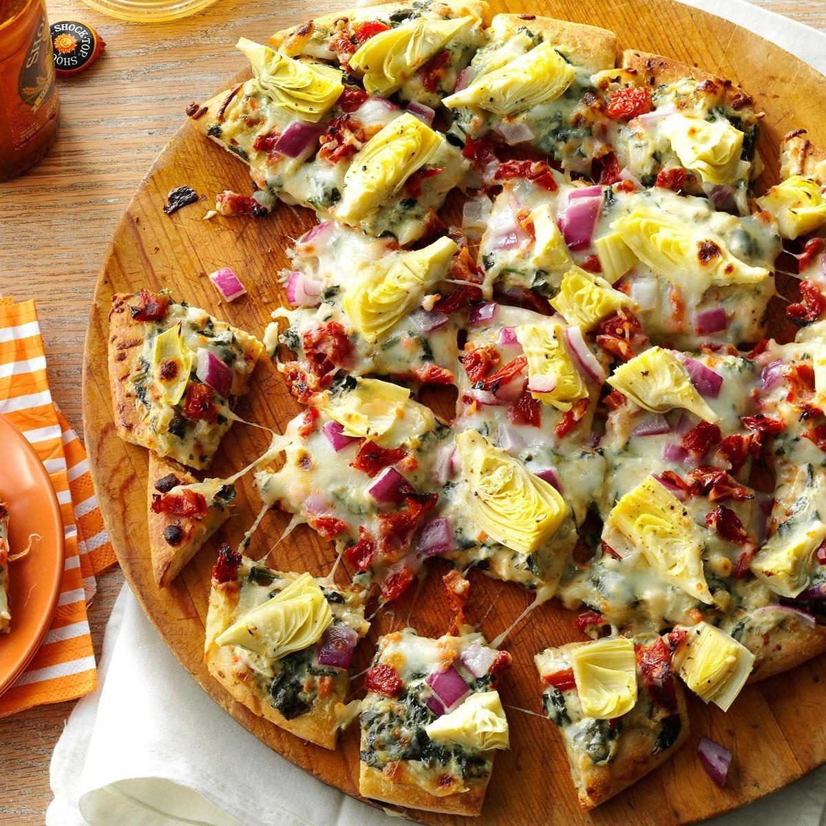 Pizza topped with cheese, artichokes, red onions, and sun-dried tomatoes, resting on a wooden board, surrounded by a rustic table setting.