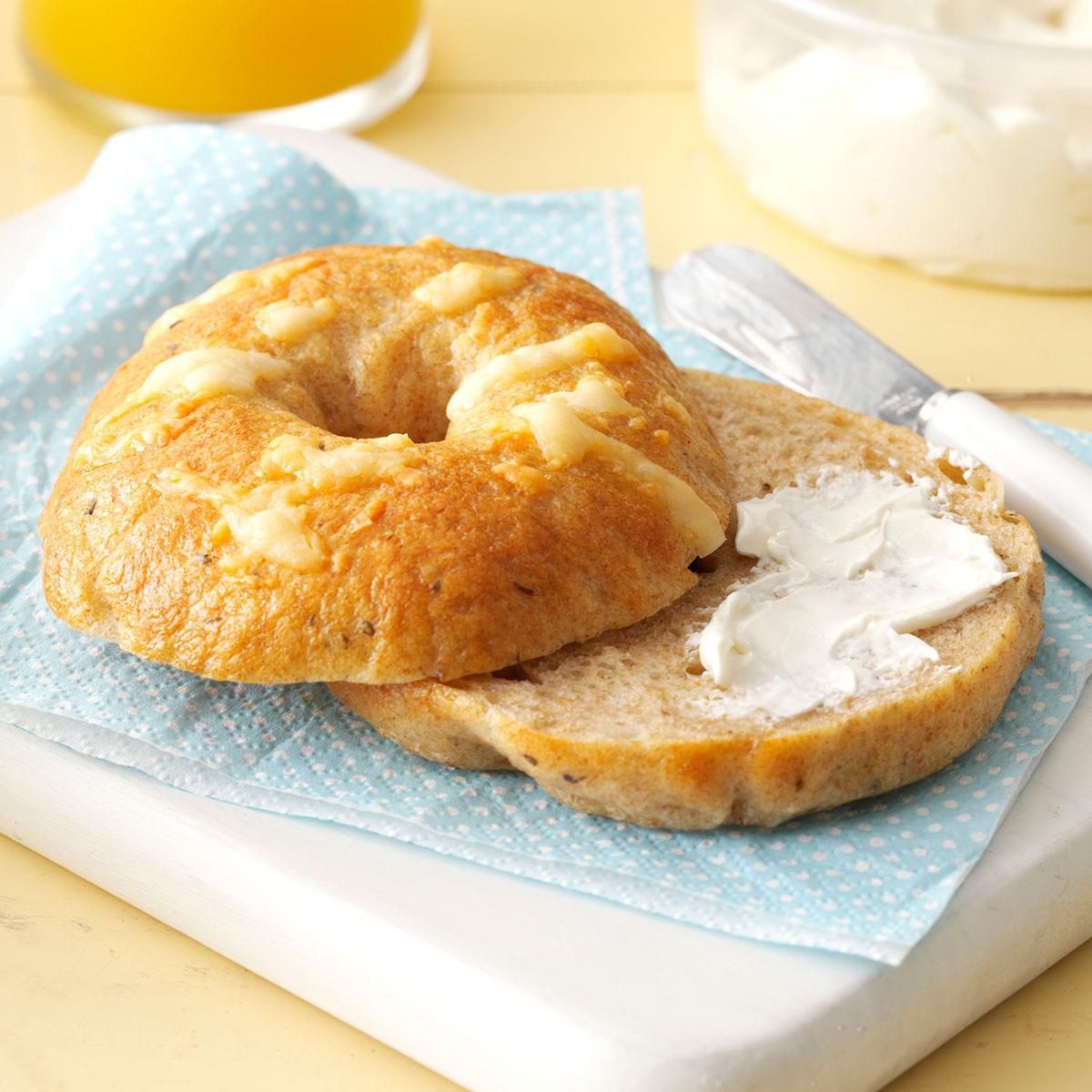 Bagel halves rest on a blue napkin; one half spread with cream cheese. A knife and orange juice are nearby.