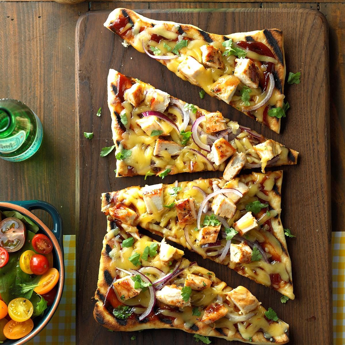 Pizza slices rest diagonally on a wooden board, topped with chicken, cheese, onions, and cilantro. A salad and beverage are nearby on the table.