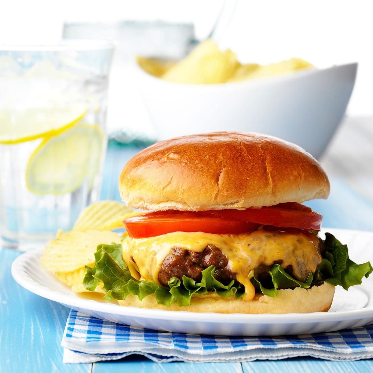 Cheeseburger resting on a blue checkered napkin, accompanied by potato chips, with a glass of lemon water in a bright dining setting.