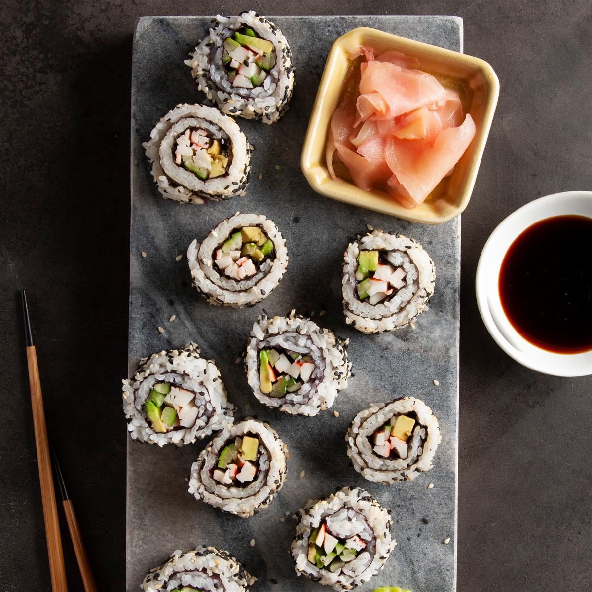 Sushi rolls with avocado and crab stick arranged on slate with pickled ginger and soy sauce, accompanied by chopsticks in a dark setting.