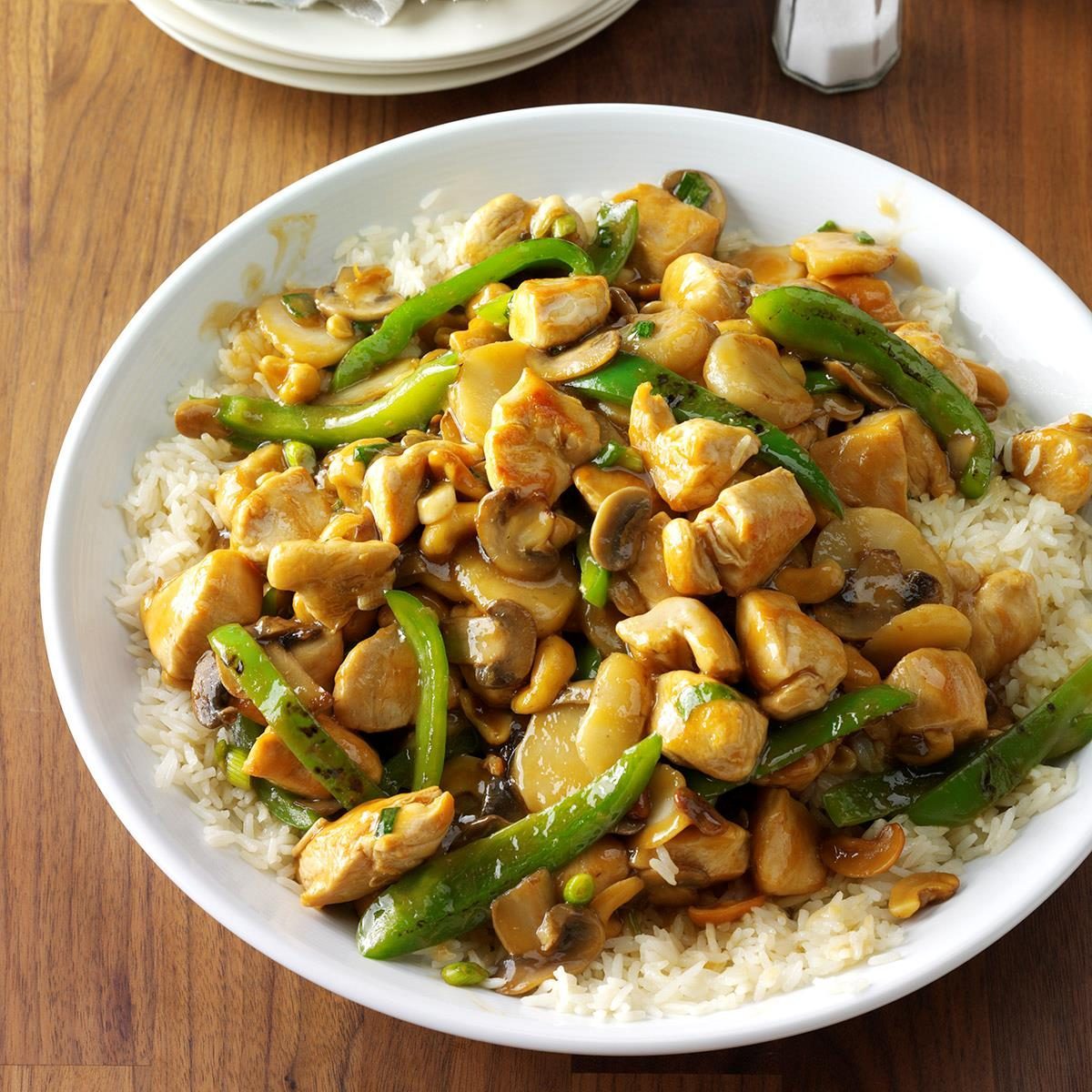 Stir-fried chicken and vegetables rest atop white rice in a white bowl, placed on a wooden table.