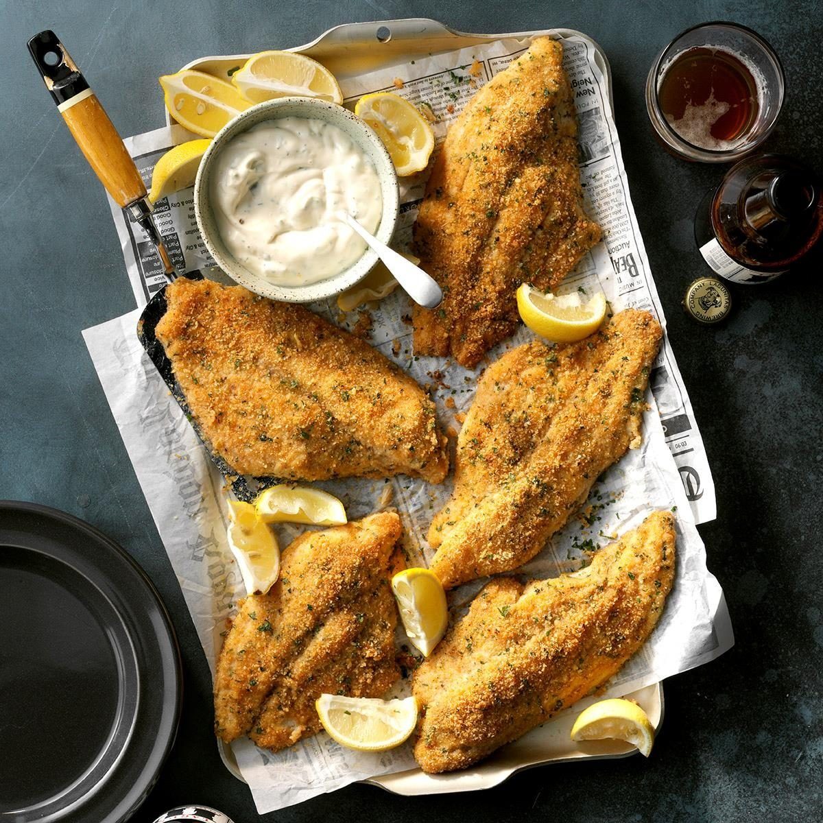Fried fish fillets rest on newspaper-lined tray, accompanied by lemon wedges and a bowl of sauce, surrounded by drinks and a dark surface.
