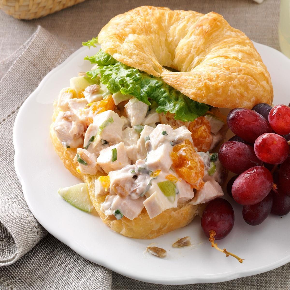 Croissant sandwich filled with chicken salad and lettuce, accompanied by a small bunch of red grapes on a white plate, placed on a linen cloth.